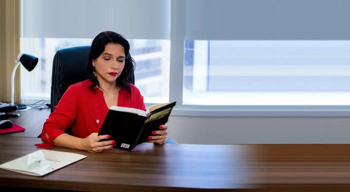 Viviane Gomes sentada à mesa do escritório, de blusa vermelha, lendo um livro diante da janela clara