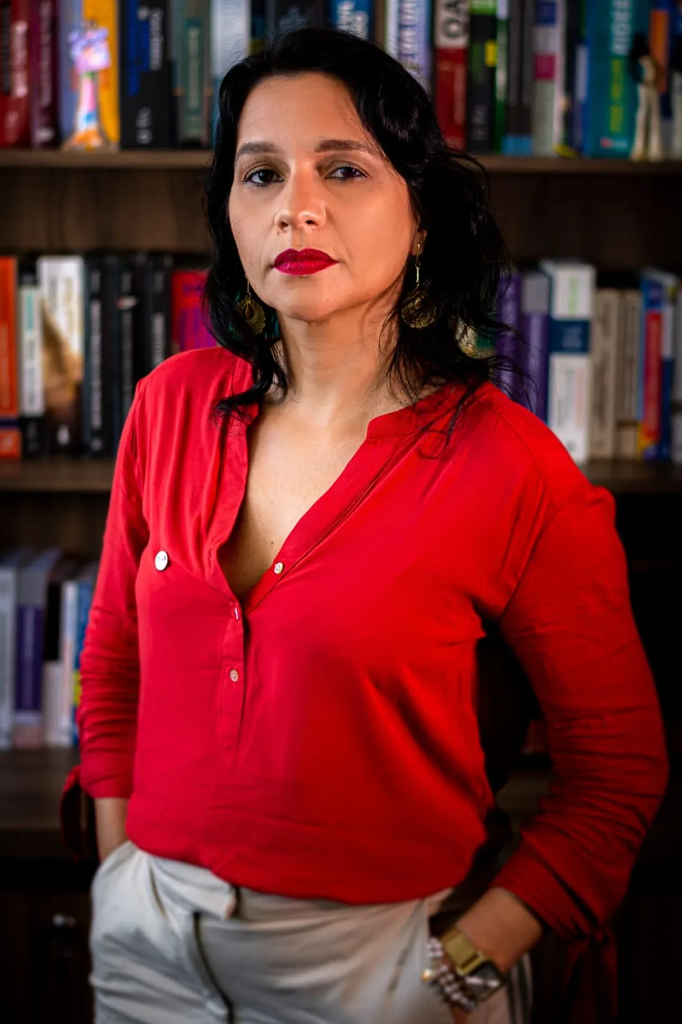 Viviane Gomes de pé diante da estante de livros do escritório, de blusa vermelha, com a mão no bolso